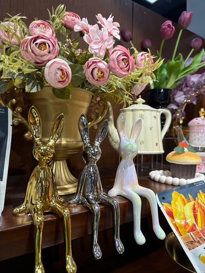 The image depicts a decorative display on a wooden shelf, likely in a shop in Phu Thuy, Vietnam. In the foreground, three ceramic rabbit figurines are perched on the edge of the shelf. From left to right, the first rabbit is gold, the second is silver, and the third is iridescent white with pastel hues. They all have long ears and dangling legs. Behind the rabbits, a large, ornate gold vase overflows with artificial pink and white camellias and other greenery. To the right of the vase, a pale yellow teapot with floral decals sits on a metal stand. Further to the right, a decorative cupcake with a red strawberry on top is placed on a white beaded mat. A package of what appears to be dried fruit, featuring bright yellow and orange slices, is also visible on the shelf, partially obscuring the bottom right corner of the image. In the background, dark wooden paneling and glimpses of other decorative items, including purple tulips and ornate pink tableware, suggest a retail environment.