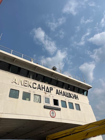 A low-angle, eye-level medium shot captures the side of a large white ferryboat against a blue sky with scattered white clouds. The ferry's name, "АЛЕКСАНДР АНАШКИН" (Aleksandr Anashkin), is prominently displayed in large, black, metallic letters. Below the name, a circular red and white sign indicates a maximum width of "40 M". Several dark-tinted square windows are visible along the side of the vessel, with some appearing to have curtains or reflections. A railing runs along the upper deck. The lower portion of the image shows part of a yellow ramp-like structure with the registration number "BC 18 01" stenciled in black. The scene is set in the city of Novoselovo, Russia.