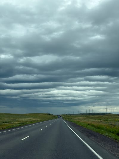 A wide asphalt road stretches into the distance under a heavily overcast, grey sky. The road has white dashed lines marking the lanes and a solid white line on the right edge. To the left of the road is a grassy embankment, and beyond that, rolling hills covered in dry grass and sparse vegetation. On the right side of the road, the terrain is also grassy with some yellow wildflowers. In the distance, several tall electrical transmission towers are visible on the horizon. The weather appears to be approaching rain, with dark, layered clouds dominating the upper portion of the frame. Two vehicles, possibly trucks or vans, are seen on the road in the far distance, heading in the same direction as the viewer. The scene is rural and appears to be located on the outskirts of Верхнеозёрное, Russia, given the typical landscape and infrastructure found in such areas.