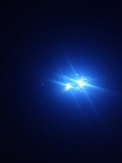 A central, concentrated bright blue light pierces the dark blue background, casting a distinct, linear reflection. The intense illumination emanates from an unseen source, creating multiple light beams that extend outwards. The overall effect is a stark contrast between the dominant light and the surrounding darkness, with some lens flare.