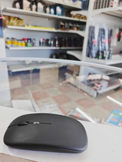 The image shows a black computer mouse placed on a white surface in the foreground. Behind the mouse, a glass counter displays various items on shelves in the background. The shelves are stocked with bottles, cans, and other products, some of which appear to be automotive fluids or cleaning supplies. The floor visible through the glass counter has a tiled pattern. The scene is likely in a store or workshop setting. There is no information in the image to suggest the location in the city of Родниковское сельское поселение, Ukraine.