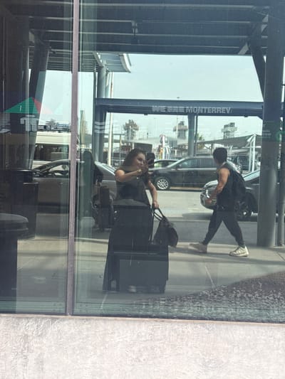 The image captures a bustling scene through a large, reflective glass window, set during a bright, sunny daytime. The overall setting appears to be a modern building, possibly an airport terminal or a similar public transit hub.
In the foreground, a woman with dark hair, wearing a dark top and watch, is visible. She holds a phone to her ear or is taking a picture, and stands beside a black rolling suitcase and a dark handbag. Her presence and luggage suggest she is a traveler waiting or passing through. Her figure appears to be inside the building, reflected and viewed through the glass. To the right, a textured, light-grey concrete wall forms part of the interior or adjacent exterior.
Beyond the glass, an outdoor area is visible. A man, wearing a dark t-shirt, dark pants, and sneakers, is walking or standing. He appears to be carrying a bag on his back. Further back, a dark-colored car, possibly a van or SUV, is parked or driving. The ground outside is paved with a textured, rocky surface.
The highly reflective nature of the glass window creates a complex visual, blending reflections of interior structures, such as dark beams and light ceilings, with the outdoor elements.
Prominently visible on a vertical structure outside the building is partially obscured text that reads "DE CIUDAD MONTERREY," clearly indicating a connection to the city of Monterrey. Another reflection shows text like "VIVA" and fragments such as "cepción" and "equipaje," which likely refer to Viva Aerobus, a Mexican airline, and airport services (reception, luggage). Given the provided context of Apodaca, Mexico, this location is strongly suggested to be Monterrey International Airport.