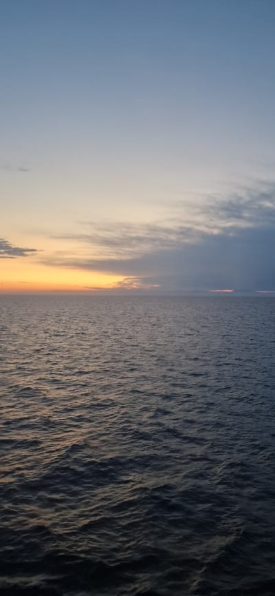 The image depicts a serene seascape at dusk. The sky transitions from a clear, pale blue at the top to hues of orange and yellow near the horizon, where scattered clouds are visible. The ocean occupies the lower half of the frame, with gentle waves reflecting the fading light. The overall impression is one of tranquility and vastness. The location is described as Unknown, Croatia, but no specific landmarks or urban elements are visible to confirm this.