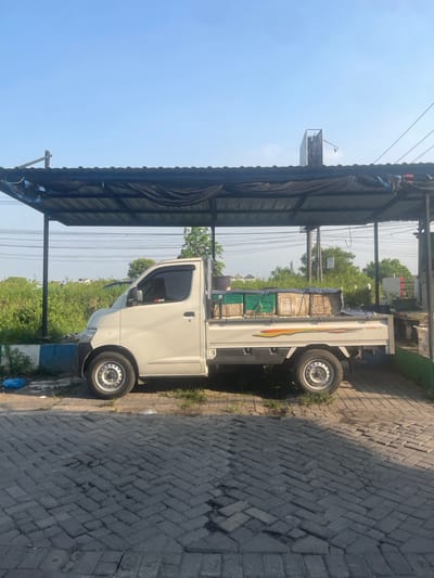 A white pickup truck is parked under a dark awning in Anggaswangi, Indonesia. The truck bed is loaded with rectangular objects, some covered in green tarps and others with beige material. The truck has a dark tinted driver's side window and a decal with red and yellow stripes on its side. The vehicle is parked on a brick-paved surface. To the left of the truck, there is a blue and white curb with some greenery growing behind it, and a blue plastic bag on the ground. The background consists of a clear blue sky, some trees, and utility poles.