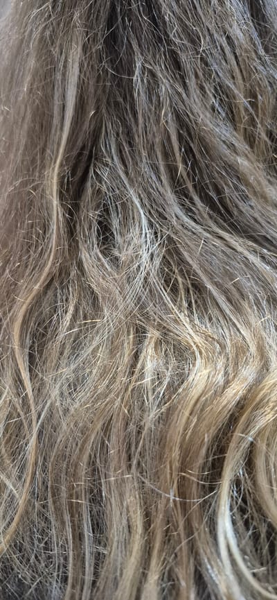 The image is a close-up, overhead shot of human hair. The hair appears to be medium to long in length and has a wavy texture. The color is a blend of natural blonde and light brown tones, with some strands appearing lighter, possibly due to highlights or sun exposure. The hair is dense and appears slightly tousled, suggesting it might be recently brushed or styled loosely. There are no other objects or discernible background elements visible in the frame. The image does not provide any information about a specific location within Póvoa de Varzim, Portugal.