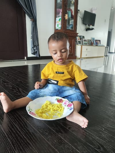 An image captured indoors in the city of Meunasahbaro, Indonesia, depicts a young child, likely a toddler, sitting barefoot on a dark brown wooden floor. The child wears a yellow long-sleeved shirt with a black patch on the chest and blue denim shorts, looking downwards with a slightly open mouth. A white plate with scalloped edges and floral patterns (red and pink roses) rests directly in front of the child, containing a pile of bright yellow rice. In the background, a dark brown door with a grey curtain is partially visible to the left. A tall, ornate dark brown wooden display cabinet with glass panels stands centrally. To its right, a low white cabinet with gold-colored trim supports multiple framed pictures, with a flat-screen television mounted on the white wall above it. A silver cylindrical object is visible against a white wall on the far right.
