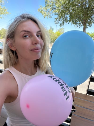 A fair-skinned woman with blonde hair pulled back is holding two balloons, one pink and one blue, in an outdoor setting. She is wearing a light pink tank top. The pink balloon is closer to the camera and has "Happy" written on it in black lettering. The blue balloon is behind the pink one and also has "Happy" written on it. Trees and a bright blue sky are visible in the background. The image appears to be taken during the daytime. The location is the city of Húmera-somosaguas-prado Del Rey, Spain.