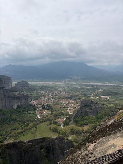 The image shows a panoramic view of Kastraki, Greece, from a high vantage point, possibly from one of the monasteries of Meteora. The foreground features rocky cliffs with vegetation clinging to the sides. In the midground, there is a town with buildings and roads winding through it. The background includes distant mountains partially obscured by clouds. The sky is overcast, creating soft, diffused lighting across the landscape.