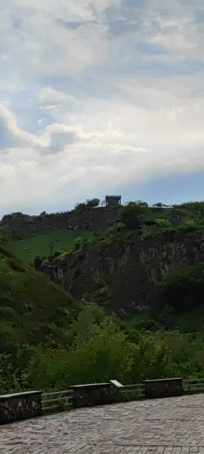 The image presents a view in Garni, Armenia, featuring a stone structure atop a ridge. The structure, possibly a small temple or monument, stands against a backdrop of a cloudy sky. The ridge is covered with sparse vegetation and some trees. Below the ridge, the landscape descends into a green valley. In the foreground, a stone-paved surface is partially visible, bordered by a stone wall and fencing.