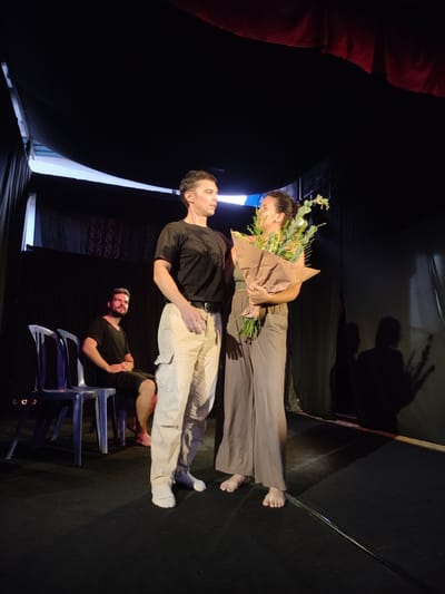 The image depicts a scene on a stage, likely in Bedulu, Indonesia, although the precise location cannot be confirmed definitively. Two people, a man and a woman, stand center stage. The man, on the left, is wearing a black t-shirt, tan cargo pants, and white socks. The woman, on the right, is holding a bouquet of greenery wrapped in brown paper and is wearing a grey tank top and grey palazzo pants. Both are barefoot.

Behind them to the left, a bearded man sits on one of two blue plastic chairs.

The background is dark, created by black curtains draped around the stage. The lighting is primarily from above, casting long shadows on the backdrop.
