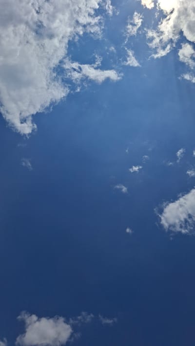 A bright, blue sky dominates the image, with scattered white cumulus clouds. The sun is partially visible in the upper right corner, casting a bright light and creating subtle sunbeams. The lower portion of the image is a deep, solid blue, with a few smaller clouds dotted throughout. The scene is a natural outdoor environment, specifically the sky above Gazalkent, Uzbekistan. There are no people, subjects, objects, actions, or interactions depicted, only the sky and clouds.