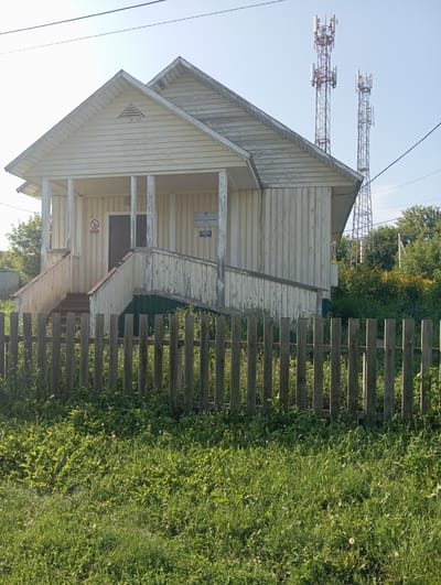 The image shows a small, weathered wooden building with a porch and steps, set behind a rustic wooden fence. The building is painted a light color, possibly white or cream, and appears to be in a state of disrepair, with peeling paint and some signs of aging. There are two cell towers visible in the background, towering over the building and surrounding trees. The area around the building is overgrown with grass and vegetation, and a few yellow flowers are visible among the greenery. A sign with a red circle and slash indicating "no smoking" is posted on the wall near the entrance. Another sign with text in Russian is also visible. The sky is clear and blue, suggesting a sunny day. The overall impression is one of a rural or semi-rural setting in Новокопылово, Russia, with a blend of older structures and modern communication infrastructure.