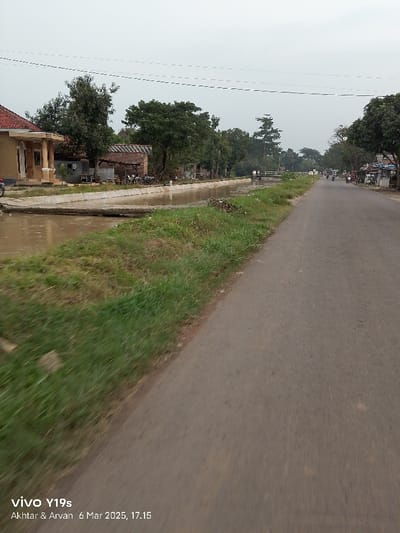 The image shows a paved road in Kecamatan Gabuswetan, Indonesia (latitude: -6.444, longitude: 108.046). There are buildings and trees on the left side of the road, and a canal with murky water running parallel to the road. The road is empty except for two figures on motorcycles in the distance. There is a small bridge crossing the canal. There is a line of power lines across the top of the image. The scene is taken during the day, with overcast skies. The photograph was taken from a moving vehicle.