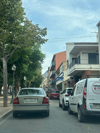 The image depicts a street scene in Torroella de Montgrí, Spain, under a sky with scattered clouds. The vantage point is from within a vehicle, looking forward down a paved road.
In the immediate foreground, the rear of a silver Volvo sedan, bearing the Spanish license plate GI 4394 BN, is centered in what appears to be a traffic lane. To the right, a white Renault Kangoo commercial van with "URAMAKI SUSHI" branding is parked along the curb, its license plate partially visible, starting with "9265 K." Behind the van, a white SUV and a red compact car are visible further down the street.
The right side of the street is lined with multi-story urban buildings. The closest visible building has a white facade, a tiled roof, a prominent balcony with a black railing, and windows with dark shutters. Further down, buildings with both white and terracotta-colored upper sections are present, some featuring balconies. Storefronts are visible along this side, with identifiable signage including "EUROMAKO," "EMPORIUM," and "CLÍNICA VETERINARIA" (Veterinary Clinic).
The left side of the street features a wide sidewalk bordered by a row of mature trees with dense green foliage, casting partial shade. Multiple streetlights with dark, decorative fixtures are interspersed among the trees. In the far background, behind the buildings on the right, the outline of mountains or large hills is discernible. No individuals are clearly visible within the frame, nor are any specific human actions or interactions evident. The scene indicates a typical urban environment with vehicular presence.