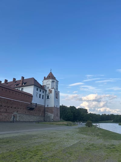 The image shows the Mir Castle Complex in the village of Mir, Grodno Region, Belarus, under a partly cloudy blue sky. The castle, a UNESCO World Heritage site, features a prominent white tower with a red, conical roof and a darker brick wall extending to the left, topped with red-tiled roofs. The castle is situated next to a calm body of water, likely a moat or lake, with a line of trees forming the background on the opposite bank. A paved area and a grassy expanse are in the foreground. Several indistinct figures are visible walking along the path by the water and near the castle entrance.