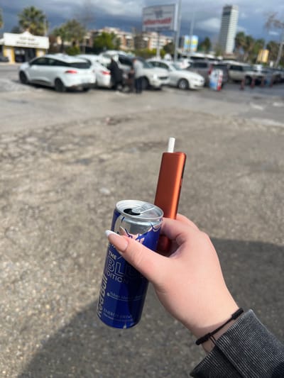 A first-person shot depicts a hand holding a blue Red Bull can and a heated tobacco product (HTP) in front of an urban scene in Bayraklı, Turkey. The hand, adorned with a bracelet, is neatly manicured with almond-shaped nails. The HTP is orange. The background reveals parked cars, trees, a tall building, and a sign, indicating a possible parking area near a commercial establishment in the city. The weather appears overcast. The ground is concrete.