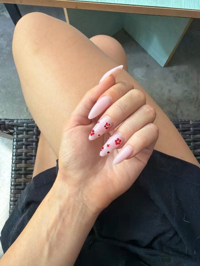 A close-up shot of a person's hand with light pink stiletto nails adorned with red glitter flower designs, resting on their thigh. The individual is wearing a black garment. The background includes dark wicker furniture and a light blue and brown table, suggesting an indoor setting or a shaded outdoor area in Prosena, Bulgaria.