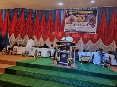 The image depicts an indoor event, likely a church service or gathering, in Ungwan Liman, Nigeria. The scene is set on a raised platform covered in artificial green turf, with a tiered arrangement of steps leading up to it. At the center of the platform stands a prominent, ornate podium adorned with gold accents and a cross. Behind the podium, a large banner with text and an image of a building is displayed against a backdrop of red and blue drapery.
Several individuals are present. A man in a light yellow garment stands at the podium, holding a microphone and speaking, appearing to be the main speaker. To his left, seated on a white couch, is another man in traditional attire. In the background, other attendees are visible seated on couches and chairs. Small tables with bottles of water are placed strategically around the seating area. The overall environment suggests a formal occasion within a church setting, characterized by its decorative elements and the presence of congregants.