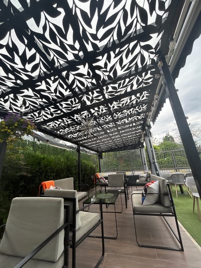 This media file depicts an outdoor seating area, likely a patio or cafe, in Dolna Banya, Bulgaria. The scene is oriented vertically, with the sky and treetops at the top. The foreground is dominated by a black, perforated metal awning with an intricate leaf pattern, casting dappled shadows. String lights are attached to the awning structure, suggesting an evening or overcast setting.
Several seating arrangements are visible. On the right side of the frame, a row of outdoor chairs with light-colored cushions and some with orange accents are arranged on a wooden deck. The chairs have dark metal frames. In the middle ground, a black metal table with a glass top is situated between two chairs. Further back, on a green, artificial turf area, several white plastic chairs are visible.
The background reveals lush green foliage, suggesting a garden or natural surroundings. The sky is overcast with some visible clouds. There are no people or animals in the image. The overall impression is one of a tranquil, shaded outdoor space, possibly designed for relaxation or dining. The time of day appears to be either late afternoon or early evening, given the lighting and the overcast sky. The location context of Dolna Banya, Bulgaria, is provided. There is no visible text in the image.