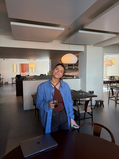 The image depicts the interior of a modern restaurant or cafe located in Marche-lez-Écaussinnes, Belgium. A female subject stands prominently in the center-left foreground, facing the viewer with a slight smile. She has dark hair, partially pulled back, and is wearing a blue plaid button-up shirt over a brown halter-neck top and dark trousers. Her left hand holds a clear glass containing a dark beverage with ice and a black straw. Her right hand, resting on a dark brown round table, holds a light blue smartphone. A closed silver-grey laptop with a visible Apple logo is positioned on the table to her left.

The background shows a spacious, well-lit dining area. To the left, a counter or bar area is visible, featuring a dark surface, various bottles, and glassware. Beyond this, additional tables and chairs are arranged. The walls are predominantly light-colored, and the ceiling features multiple rectangular white acoustic panels suspended below the main ceiling. Several distinctive pendant lights illuminate the space; one large, white, ribbed spherical lamp hangs directly above the bar area, and other modern, rounded lamps are visible further in the background. Seating includes dark wooden chairs with light-colored woven seats and small dark wooden stools. The floor is a dark, smooth surface. Artwork, including a large rectangular piece with reddish-brown tones, is visible on a back wall. The overall environment suggests a contemporary and minimalist design.