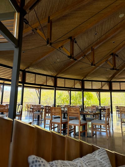 The image displays an empty dining area within a building in La Fortuna, Costa Rica. The space features a high, sloped, multi-faceted ceiling constructed from exposed dark wooden beams and metal tension rods, with light-brown woven paneling. Recessed circular lighting fixtures are integrated into the ceiling. The perimeter of the room is defined by large, open window frames offering panoramic views of dense, vibrant green foliage. Above these main window sections, horizontal wooden slat panels are visible. The floor is dark and reflective. Numerous dark-topped square dining tables are uniformly arranged throughout the area, each accompanied by wooden chairs with light upholstered seats. Tables are pre-set with glassware and small white plates. In the foreground, a wooden partition with a white patterned cushion is partially visible. No individuals are present.