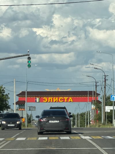 The image captures a street scene in Russia. The perspective is from a vehicle moving along a road, indicated by the presence of lane markings and other cars. The prominent subject is a large, red building with yellow lettering that reads "ЭЛИСТА." The building appears to be a commercial establishment, possibly a store or a business complex.
Several cars are visible on the road. One black car is parked in the foreground, and another black car is in the lane ahead, with its license plate partially visible as "B819XC 09". Further down the road, another black car with license plate "E545CH" is approaching. A traffic light showing a green signal is also present, suggesting the vehicles are at an intersection.
The sky is filled with dramatic, cloudy formations, suggesting a dynamic weather pattern, possibly overcast or transitioning. Streetlights and overhead wires are strung across the scene, typical of an urban or suburban environment. In the background, trees and other structures can be seen, contributing to the overall setting of a developed area. The time of day appears to be daytime, given the lighting conditions. No people are clearly discernible in the image. The overall impression is a snapshot of everyday life on a Russian road.