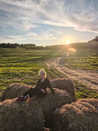 A wide shot captures a rural landscape at sunset in what appears to be S стеклянный, Russia. The warm, golden sun is low on the horizon, partially obscured by distant trees and casting a soft glow across the scene. The sky above features light blue hues with wispy, streaky white clouds.

In the foreground, a woman with blonde hair styled in a bun is seated comfortably on several large hay bales. She wears dark clothing and distinctive multi-colored, striped socks with dark red sneakers. Her head is tilted downwards, and she appears relaxed or contemplative, bathed in the warm sunset light.

Behind her, a vast green field stretches into the distance, bordered by a dense line of forest under the setting sun. A winding dirt road cuts through the field, and in the mid-ground, a dark-colored car is visible driving along this road, kicking up a trail of dust that is illuminated by the strong backlight of the sun. The overall atmosphere is peaceful and rustic, highlighted by the tranquil golden hour lighting. There is no visible text in the image.