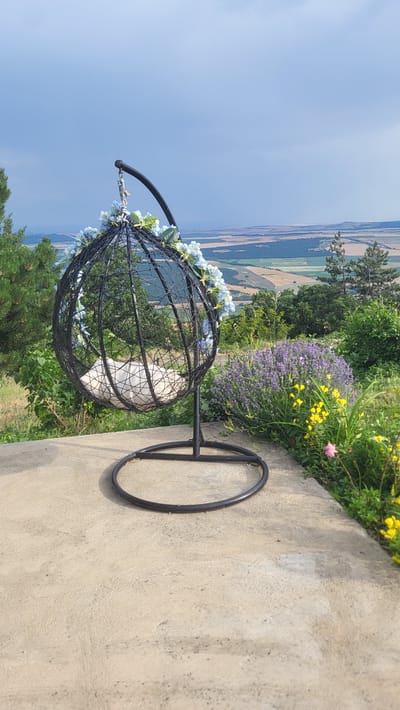 This media file features an outdoor scene in Topolitsa, Bulgaria. The primary subject is a black, wire-frame hanging chair, adorned with light blue artificial flowers around its rim. The chair is suspended from a metal stand with a circular base. Inside the chair, a white cushion is visible.
The chair is positioned on a concrete pathway that runs alongside a garden area. Behind the chair, a vibrant garden bursts with greenery, featuring a dense patch of purple lavender flowers and smaller yellow flowers, with a single pink flower also present. In the background, a sweeping landscape unfolds, showing rolling fields and distant hills under a clear blue sky. The perspective suggests the location is elevated, offering a panoramic view.
The lighting indicates it is daytime, likely mid-morning or afternoon, with bright sunlight illuminating the scene and casting subtle shadows. The weather appears clear and pleasant, conducive to outdoor activities. There is no visible text in the image. The overall impression is one of tranquility and natural beauty, inviting relaxation and enjoyment of the scenic surroundings.