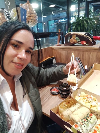 The image depicts an adult female seated at a wooden table inside what appears to be a casual dining establishment. She is positioned slightly to the left, facing the camera with a smile. She has dark hair and is wearing a white collared shirt under a dark green padded jacket. Her left hand, holding a pair of white chopsticks, grips a piece of fried or breaded sushi.
On the wooden table to her right are two open cardboard takeout boxes containing various sushi rolls, including some with fried coatings, and a section with what appears to be chips or crisps topped with white and red elements. A small clear dish containing a red sauce and a stack of three small, dark, circular dishes are also on the table. Behind these items, pens and a black stapler are visible. A digital timestamp "24/02/2026 17:40" is overlaid in the lower right corner.
The background features a wooden counter or shelf decorated with a wooden wine rack holding a red wine bottle, a wooden cross-shaped ornament, and a large glass container filled with wine corks. Wine bottles are also visible. A large glass partition separates the dining area from an outer space featuring green foliage and ambient lighting, indicating either an exterior view or another section of the interior. The image does not contain explicit identifiers for the city of Tumbaco, Ecuador.