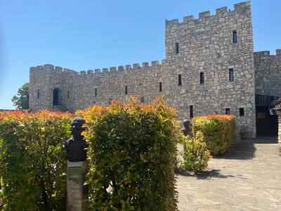 The image shows a stone castle with crenellated walls and small windows against a clear blue sky. In the foreground, there are neatly trimmed bushes with reddish-brown leaves and a bust of a man on a stone pedestal. The bust has a plaque attached to it, but the text is too small to read clearly. The ground is paved with large, irregular stones. To the right, a dark entrance to a building is visible, and a sign with Cyrillic lettering hangs above it. The scene appears to be an outdoor historical site or museum, likely in Pliska, Bulgaria, given the architectural style.