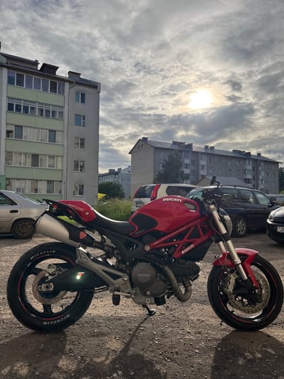 A red Ducati motorcycle is parked on a gravel surface in front of apartment buildings in Vileyka, Belarus. The sun is visible through the clouds, casting shadows on the ground. The motorcycle is the primary subject, with its brand name prominently displayed. Several cars are parked in the background, and one building on the left has a green facade. The overall scene depicts an urban environment on a cloudy day.
