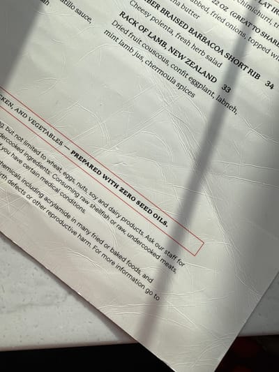 The image displays a close-up, angled view of a white restaurant menu, likely captured indoors. A significant diagonal shadow cuts across the top and middle sections of the menu.
The menu lists several dishes, including "BER BRAISED BARBACOA SHORT RIB" for 34, described with "Cheesy polenta, fresh herb salad." Another item, "RACK OF LAMB, NEW ZEALAND," is priced at 33 and accompanied by "Dried fruit couscous, confit eggplant, labneh, mint lamb jus, chermoula spices." Other partially visible menu entries mention "chimichurri," "fried onions," and "butter."
A key section, highlighted by red lines, declares "CKEN, AND VEGETABLES — PREPARED WITH ZERO SEED OILS." Below this, a comprehensive consumer advisory is present. It states that items may contain allergens "not limited to wheat, eggs, nuts, soy and dairy products" and advises patrons to "Ask our staff" for information, especially if they "have certain medical conditions." The advisory also warns about the risks of "Consuming raw shellfish or raw, undercooked meats." Additionally, a warning about "chemicals including acrylamide in many fried or baked foods" that can cause "birth defects or other reproductive harm" is visible, directing users to a source for more information.
The menu material appears to be a textured, off-white paper or synthetic material, with some minor creases visible across its surface. The overall impression is that of a detailed and informative restaurant menu from a dining establishment in Solana Beach, USA.