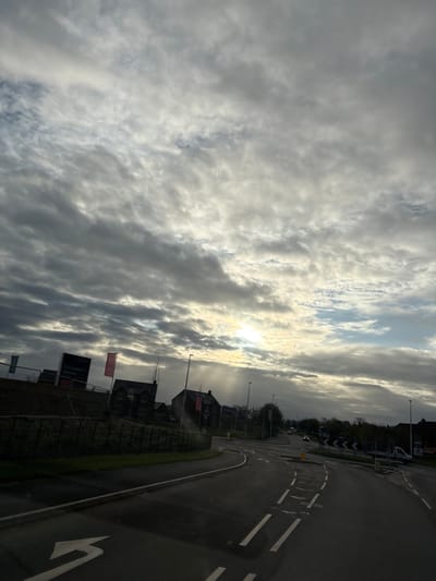 The image captures a view in Oswestry, United Kingdom, featuring a street and cloudy sky. The street, marked with painted road lines and arrows, suggests active traffic flow. Adjacent to the road is a building and perimeter fencing with signage. Overhead, the sky is dominated by thick, layered clouds with some sunlight visible through the gaps.