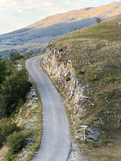 The image shows a winding asphalt road in a mountainous, rural landscape. The road curves to the right, flanked by a steep, rocky incline on the right and dense green foliage on the left. In the background, rolling hills covered in dry grass and sparse trees stretch towards the horizon under a cloudy sky. A wooden signpost with two directional arrows is visible on the right side of the road. The text on the sign appears to be "VIDIKOVAC DUBCEVICI". This scene is consistent with the mountainous terrain surrounding Bukovac, Montenegro, known for its scenic roads and natural beauty.
