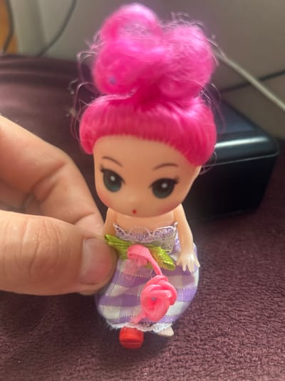 A close-up, eye-level, slightly angled indoor shot shows a hand holding a small doll. The doll has bright pink hair styled in two buns, large green eyes, and is wearing a purple and white checkered dress with a lace trim. A pink rose decoration is attached to the front of the dress. The background is blurred, showing a dark surface and a black electronic device. The image does not contain any information about the location in the city of Супонево, Russia.