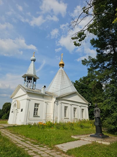 The image shows the Church of the Resurrection of Christ, located in the village of Ulitkino, Moscow Oblast, Russia. The white church has a bell tower and a golden dome. In front of the church is a monument, a bust of a woman on a black pedestal, surrounded by grass and flowers. A paved path leads up to the church. The sky is blue with white clouds, and trees are visible in the background.