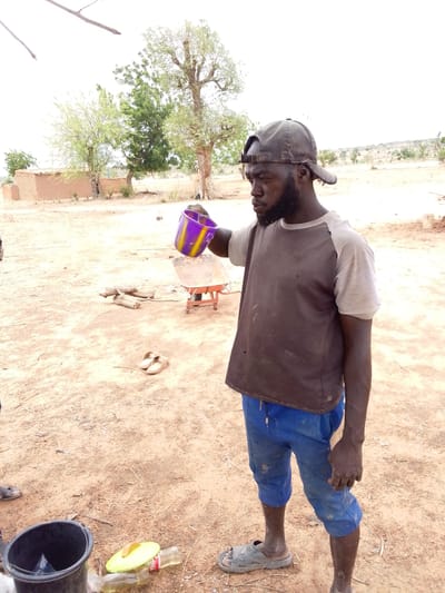 The image depicts a man in Mulke Barikin Sayejo, Nigeria, standing outdoors in a dry, dirt-covered area. He is dark-skinned, bearded, and wearing a dirty baseball cap, a two-tone t-shirt (dark brown torso, light gray sleeves), blue cropped pants, and sandals. He is holding a purple and yellow plastic cup, tilting it as if pouring liquid. Behind him is a small red wheelbarrow and some chopped firewood. In the background, there are sparse trees, a small mud structure, and dry terrain. On the ground near the man's feet is a black bucket, a plastic bottle, and discarded items. The overall scene appears arid and rural.