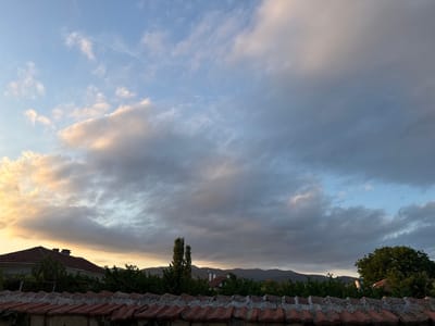 This image captures a rural landscape in Blatets, Bulgaria, during what appears to be either sunrise or sunset. The sky is filled with dramatic, textured clouds, transitioning from warm golden hues on the left to cooler grays and blues on the right. These clouds suggest a dynamic weather pattern, perhaps a transition from fair to unsettled conditions, or vice-versa.
The foreground features a rustic stone wall topped with red roof tiles, some of which are slightly weathered and covered in moss, indicating an established and potentially aged structure. Behind the wall, a low-lying settlement is visible. A house with a red tiled roof and a chimney is prominent on the left. Further back, a dense line of green trees forms a natural barrier, behind which rolling hills or mountains form the distant horizon. Two tall, slender trees stand out against the skyline, flanking a smaller building nestled amongst the foliage. The overall scene evokes a sense of peaceful, rural tranquility, with the dramatic sky adding a touch of natural grandeur. The time of day is suggested by the warm light illuminating the clouds on the left, contrasting with the darker, more shadowed areas on the right.