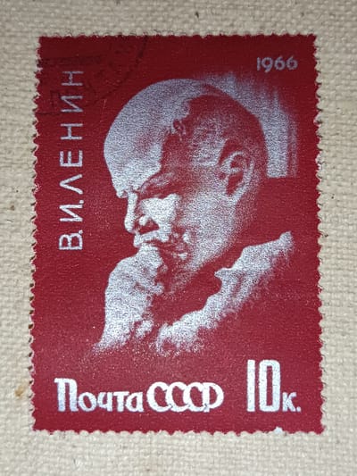 This is a close-up photograph of a red postage stamp featuring a silver-toned image of Vladimir Lenin. The stamp is printed with Cyrillic text. Vertically along the left edge, the text reads "В.И. ЛЕНИН" (V.I. Lenin). In the upper right corner, the year "1966" is printed. Along the bottom edge, the text reads "Почта СССР 10к." (Post of the USSR 10 kopecks). The stamp has a perforated edge. The background of the image appears to be a textured beige surface, possibly linen or burlap. There is no information within the image or its description that pertains to the city of Onaldy, Turkmenistan.