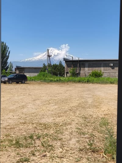 The image depicts an outdoor scene with Mount Ararat in the background, partially obscured by clouds and smoke. The foreground shows a dirt and dry grass field. To the left, a black SUV is parked. In the mid-ground, there are low-rise buildings constructed with grey bricks and metal roofs. Vegetation, including trees and bushes, is visible between the buildings and in front of them. A wooden utility pole with wires stands near the center of the image. The sky is clear and blue. The scene appears to be in a rural or semi-urban area, likely in or around Berkanush, Armenia, given the presence of Mount Ararat.