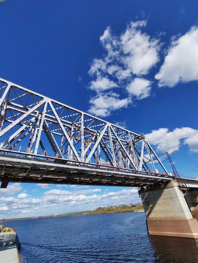 A low-angle, wide shot captures a steel truss bridge crossing a wide body of water, likely a river, under a bright blue sky dotted with fluffy white clouds. The bridge's steel structure is complex, with diagonal, vertical, and horizontal elements forming triangular patterns. Some parts of the steel are painted white, while others appear to have a rusty, reddish-brown hue. The bridge deck appears to be made of steel as well.
The bridge is supported by a massive concrete pier that emerges from the water. The pier is wider at its base and tapers upwards. The water is dark blue with visible ripples and small waves. A shoreline with trees and buildings is visible in the distance under the bridge.
In the lower left corner of the image, part of a barge is visible, suggesting the photograph was taken from a boat. A power line or some sort of cable hangs down from the bridge support structure on the right.
The overall environment is bright and sunny, suggesting a clear day. The image is well-composed and captures the grandeur of the bridge and its surroundings. There is nothing in the image to confirm that the location is in Tarkanovo, Russia.
