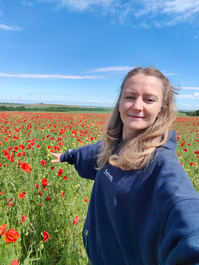A selfie shows a woman standing in a field of red poppies under a blue sky with scattered clouds. She is fair-skinned with blonde hair and is wearing a dark blue hooded sweatshirt. She is smiling slightly and extending one arm outwards towards the flowers. The field stretches into the distance, with a line of trees and green hills on the horizon. Based on the landscape, it is unlikely the location is in or near Зеленогорское сельское поселение, Ukraine, since this area is generally wooded and near the coast. Зеленогорское сельское поселение is a region known more for forest landscapes than large open poppy fields. The scene appears to be a rural setting.