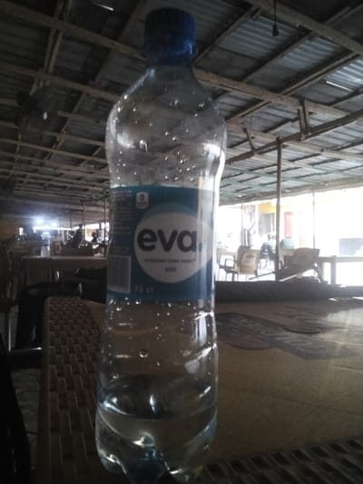 An Eva water bottle stands on a woven table. The bottle is mostly full, with condensation droplets clinging to its surface. The label indicates it's a "premium table water" that is "still" and contains 75 cl. Behind the bottle, the scene opens to a semi-outdoor dining area. Tables and chairs are visible under a corrugated metal roof, supported by wooden beams. The lighting suggests daytime, possibly under shade.
