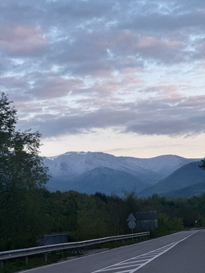 The image captures a serene landscape featuring a mountain range under a cloudy sky, with a portion of a highway in the foreground. The photograph appears to be taken during the golden hour, either at dawn or dusk, judging by the soft, diffused light and the delicate pink and orange hues visible within the cloud formations against a predominantly blue-gray sky.
In the mid-ground, a majestic mountain range dominates the horizon. The slopes of the closer mountains are covered in dense, dark green trees and foliage. Behind these, further peaks rise, appearing lighter in color, possibly indicative of snow cover or strong illumination from the low sun, and exhibiting a soft, muted blue-gray tone characteristic of atmospheric perspective.
The foreground on the right side of the image shows a section of an asphalt highway or road. It features distinct white lane markings, including a solid line along the edge and several dashed lines for traffic lanes. A metal guardrail runs parallel to the road. Beyond the guardrail and the road, a sloped area is densely covered with dark green trees and vegetation. Partially obscured within the foliage, a dark, rectangular structure, possibly a sign or a building, is faintly visible, along with a small, indistinct triangular road sign.
No discernible text is visible on any signs or objects. The overall scene conveys a sense of calm and natural beauty, characteristic of a mountainous region, in this case, Mezdreya, Bulgaria.