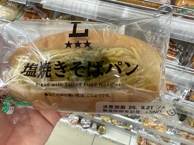 A human hand with light pigmentation holds a clear plastic-wrapped "Salted Yakisoba Pan" (bread with salted fried noodles) in a retail store aisle in the city of Chūō, Japan. The elongated, light-brown bread bun is split and filled with yellowish stir-fried noodles, topped with green flakes, likely dried herbs. The packaging displays a stylized "L" logo with three stars, Japanese text "塩焼きそばパン" (Salted Yakisoba Pan), and the English translation "Bread with Salted Fried Noodles." Smaller Japanese text indicates that black grains in the salted sauce are pepper. A visible consumption deadline reads "消費期限 25. 9. 21 /A" (September 21, 2025, assuming Reiwa 7) and a manufacturing plant code "+SNO." The background is a blurred view of metal shelves stocked with various other individually packaged baked goods and snacks, including an item labeled "ベーグルクリームチーズ" (Bagel Cream Cheese).