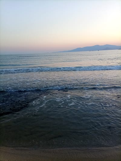 The image depicts a serene seascape at dusk near Vivlos, Greece. The sky transitions from a pale blue at the zenith to soft hues of pink and orange near the horizon, where a distant mountain range is silhouetted. The sea is calm, with gentle waves breaking on a sandy beach in the foreground. The water is a deep blue-green, reflecting the fading light. The beach appears wet and smooth near the water's edge, with darker, possibly rocky, areas visible beneath the shallow water. No people or man-made objects are present in the scene.