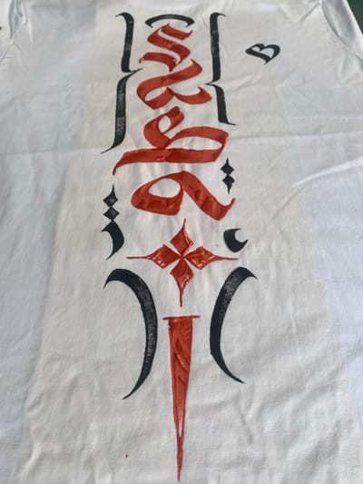 On a white cloth, likely a shirt, a decorative design is painted in black and orange. The design appears to be inspired by Tibetan or similar calligraphic styles, featuring stylized characters and symbols. A small heart-like shape is visible at the top left. The cloth is wrinkled. The photograph was taken at Baan Nai Suan, Thailand.