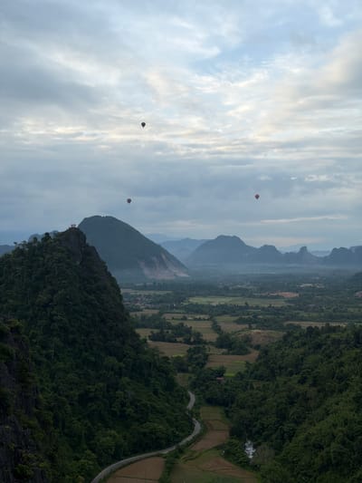 The image captures a panoramic view of a valley in Naka, Laos, dominated by lush green mountains and a cloudy sky. Several hot air balloons are visible in the sky, adding a sense of adventure to the serene landscape. In the foreground, a steep, tree-covered mountain on the left provides a dramatic contrast to the rolling hills and flat plains in the distance. A winding road snakes through the valley, leading to small villages nestled amongst the fields. The overall atmosphere is peaceful and idyllic, with the hot air balloons suggesting a tourist activity.