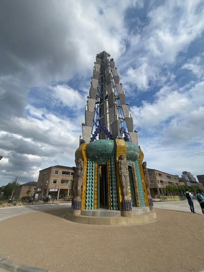 This is a vertical, eye-level shot of a tall, cylindrical sculpture adorned with colorful mosaic tiles and metallic elements, set against a backdrop of a cloudy sky. The sculpture has a wide, decorative base made of carved stone and shimmering turquoise, yellow, and blue tiles. Rising from the base is a sleek, metallic structure with numerous angled panels, resembling a finial or a stylized plant. The sculpture is situated in an urban or park-like setting with modern buildings visible in the background. The ground in front of the sculpture is paved with a sandy-colored material. A few people are present in the distance, walking and appearing to engage in everyday activities. The clouds suggest it might be daytime, with a mix of sun and shade. Signs are visible on one of the buildings, including "REMA 1000," indicating a commercial establishment. The overall impression is one of modern public art within a contemporary urban environment.