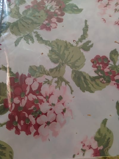 The image is a close-up of a floral pattern, likely wallpaper or fabric, with a light-colored background. The pattern features clusters of flowers in shades of pink and deep red, interspersed with green leaves and stems. Some small, dark specks, possibly debris or imperfections in the material, are visible on the surface. The image is well-lit, showcasing the detail and texture of the floral design. The photo appears to be taken indoors, possibly in a room or a studio, but no specific location in the city of Lingiai, Lithuania, can be identified. There are no people or discernible actions taking place.